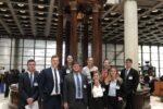 Young adults in professional attire stand before a historic bell in Lloyds of London