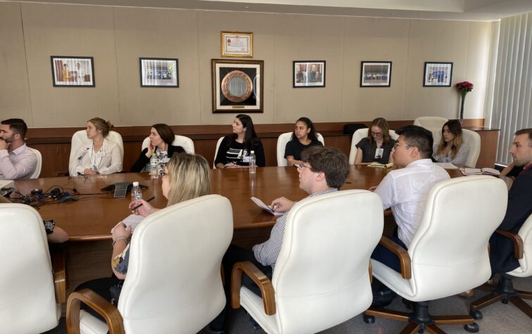 Young adults in a boardroom seated at an oval wooden table listen and take notes during a presentation