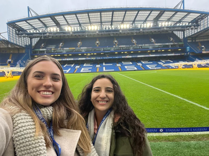 Two young women standing by the pitch at Chelsea Football Club.