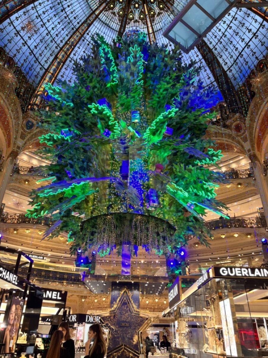 Green, blue, and purple crystal chandelier hanging beneath a glass dome in a multi-story shopping center