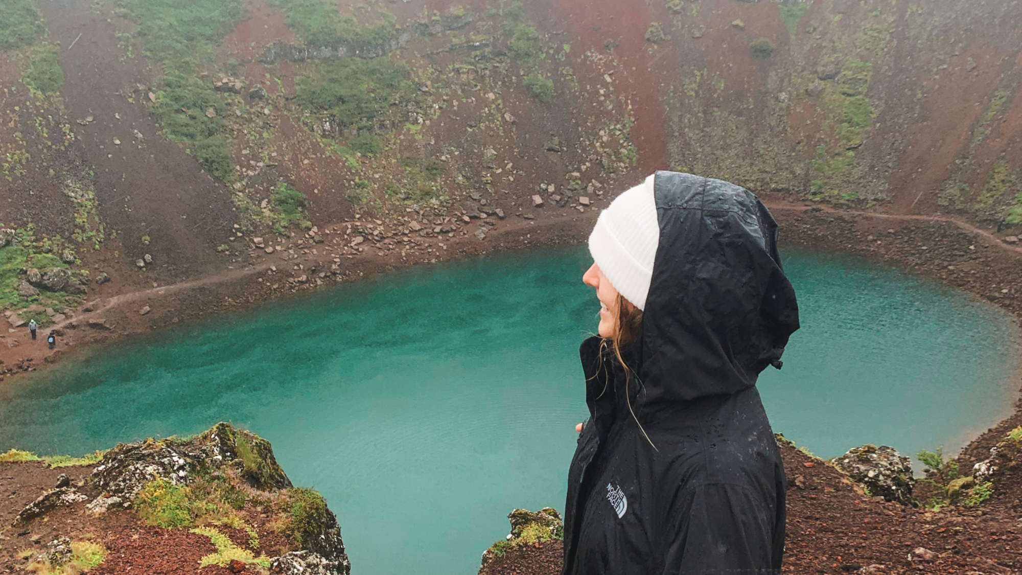 A young woman stands above a water filled crater in Iceland.