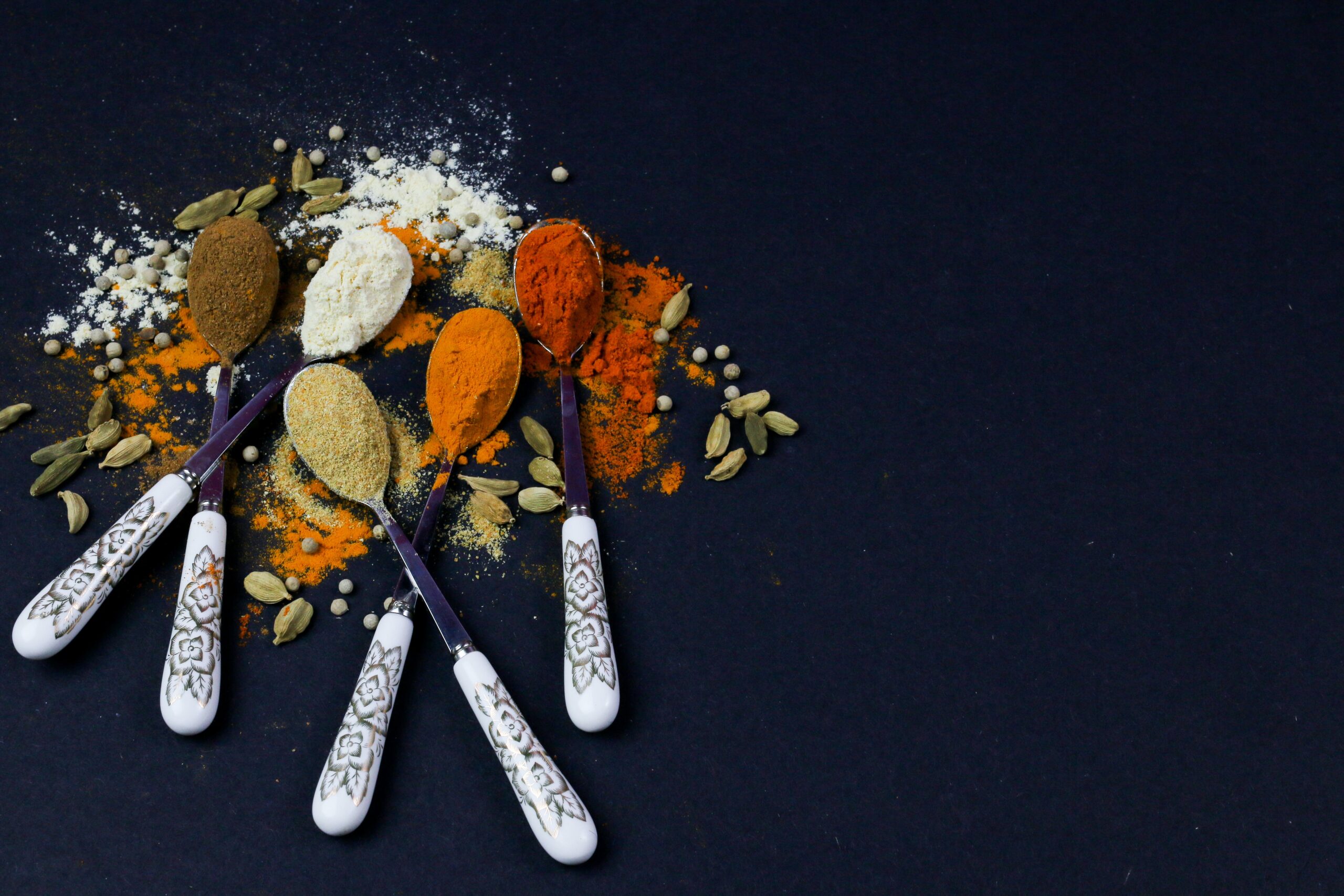 Five spoons holding a variety of spices resting on a dark tabletop surface.
